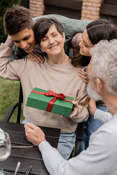 Smiling middle aged woman holding gift box and hugging children near blurred husband during picnic and parents day celebration at backyard in june, celebrating parenthood day concept - Φωτογραφία, εικόνα