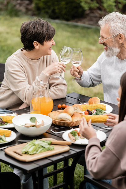BBQパーティー中の夏の食べ物や十代の娘の近くでワインの中年の妻と夫の乾杯グラスと裏庭での親の日のお祝い、家族の絆の概念を大切に - 写真・画像