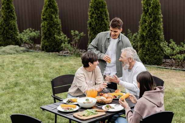 Emocionados e alegres pais de meia-idade brindando com copos de vinho perto de crianças e comida de verão durante a festa do churrasco no quintal, valorizando o conceito de laços familiares, passando tempo juntos - Foto, Imagem