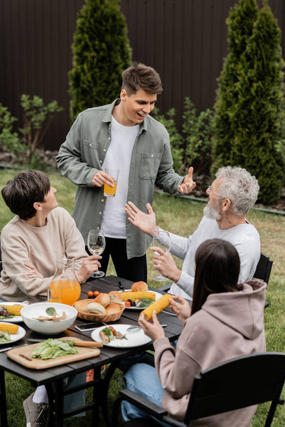 Młody i uśmiechnięty mężczyzna trzyma sok pomarańczowy i rozmawia z ojcem w średnim wieku w pobliżu rodziny podczas imprezy bbq i uroczystości dzień rodziców na podwórku, pielęgnując koncepcję obligacji rodzinnych - Zdjęcie, obraz