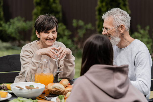 Cheerful middle aged woman holding wine glass and siting near blurred daughter and husband during summer bbq party and parents day celebration at backyard, cherishing family bonds concept - Photo, Image