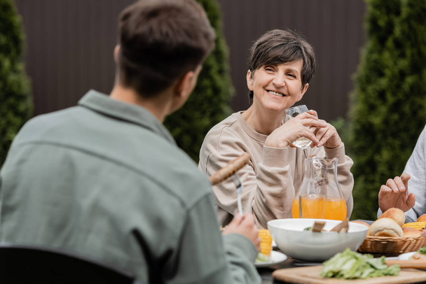 Uśmiechnięta kobieta w średnim wieku trzymająca kieliszek wina i patrząca na rozmazanego syna w pobliżu męża i letniego jedzenia podczas uroczystości Dnia Rodzica na podwórku, pielęgnująca koncepcję więzi rodzinnych - Zdjęcie, obraz