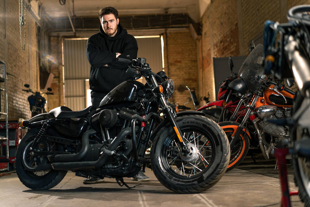クロスアームでオートバイの近くに立っている実際のオートバイのワークショップガレージで黒いフーディーの深刻なひげ付きバイカー - 写真・画像