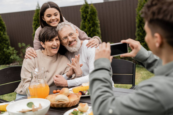Lächelndes Teenager-Mädchen umarmt Eltern mittleren Alters in der Nähe verschwommenen Bruder, der Foto auf dem Smartphone in der Nähe von BBQ-Essen während der Eltern-Tag-Feier im Hinterhof, besonderen Tag für Eltern Konzept - Foto, Bild