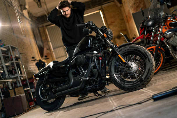 upset bearded biker in black hoodie in motorcycle workshop garage standing near motorcycle holding head - Fotó, kép