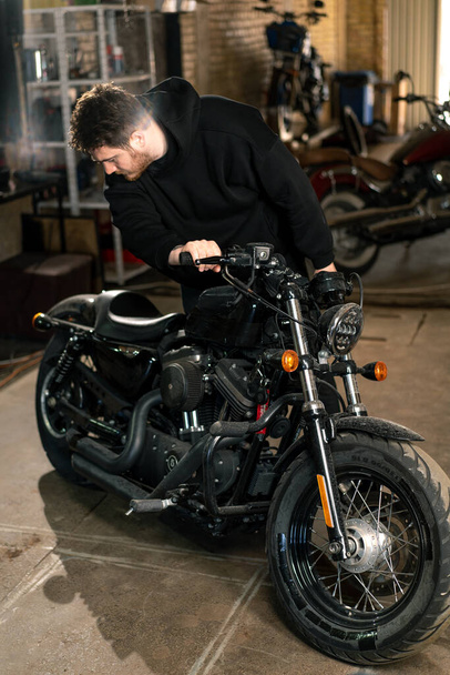 serious bearded biker in black hoodie in motorcycle workshop standing near motorcycle inspecting it for repair - Photo, Image