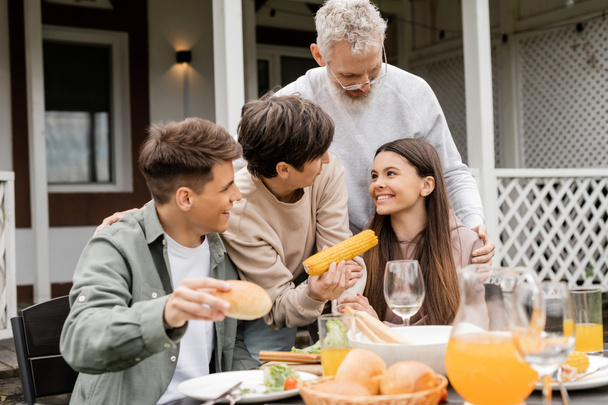 Uśmiechnięci rodzice w średnim wieku przytulający nastoletnią córkę w pobliżu syna i smaczne jedzenie podczas imprezy bbq i uroczystości dnia rodziców na podwórku w czerwcu, jakość czasu z koncepcją rodziców  - Zdjęcie, obraz