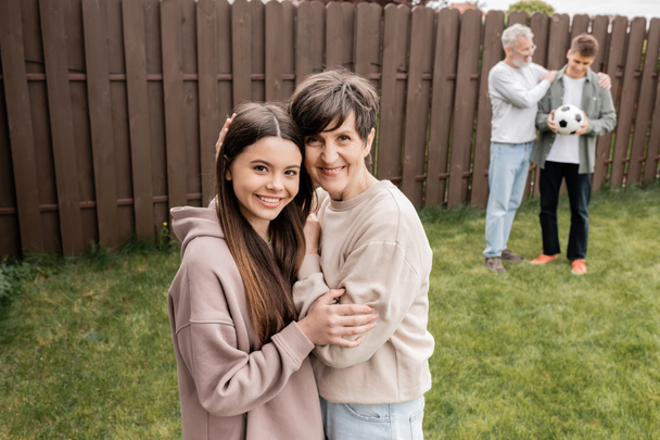 Smiling middle aged woman hugging teenage daughter and looking at camera near blurred family with football during parents day celebration at backyard, quality time with parents concept  - Photo, Image