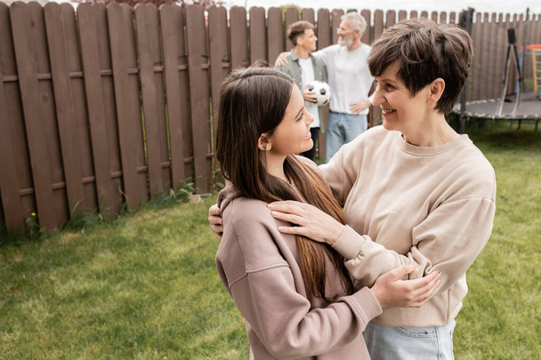 Positive teenage girl hugging middle aged mom near blurred family with football during parents day celebration at backyard in june, quality time with parents concept, tradition and celebration - Photo, Image