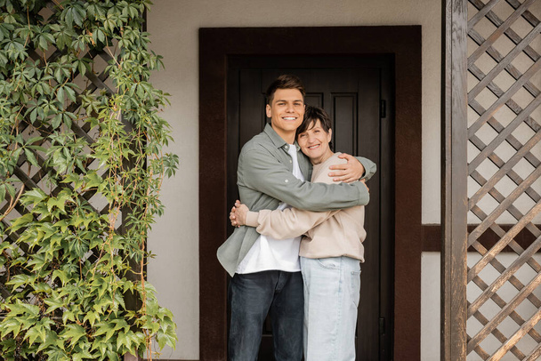 Веселый молодой человек обнимает мать средних лет и смотрит в камеру, стоя вместе на крыльце дома во время празднования дня родителей, качественное время с родителями концепции  - Фото, изображение