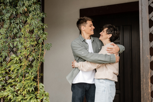 Side view of positive young man hugging middle aged mother while celebrating parents day and standing on porch of house in june, quality time with parents concept, special occasion - 写真・画像