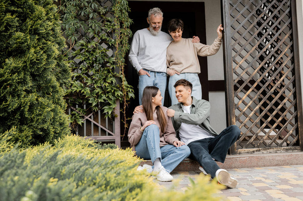 Cheerful middle aged parents hugging and looking at children sitting on porch of house during parents day celebration at backyard, family traditions and celebrations concept - Photo, Image