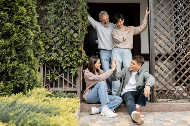 Positive middle aged parents looking at children giving high five and standing on porch of house during parents day celebration in june, family traditions and celebrations concept, special occasion - Photo, Image