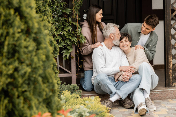 子供の近くの家のポーチに座っている間に手を握っている中年の両親を笑顔にし、裏庭で両親の日を祝う、家族の伝統とお祝いのコンセプト、特別な機会 - 写真・画像