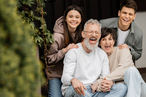Positive children looking at camera while hugging middle aged parents holding hands and sitting on porch of house during parents day celebrating , parent-child relationship concept - Photo, Image