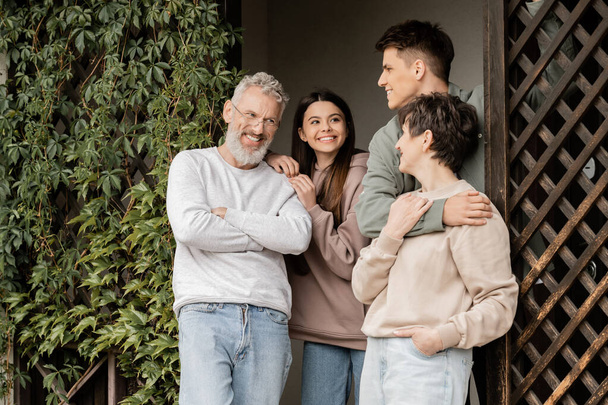 Vrolijke jonge man knuffelen van middelbare leeftijd moeder in de buurt van vader en tienerzus terwijl staan op de veranda van het huis en vieren ouders dag in juni, ouder-kind relatie concept - Foto, afbeelding