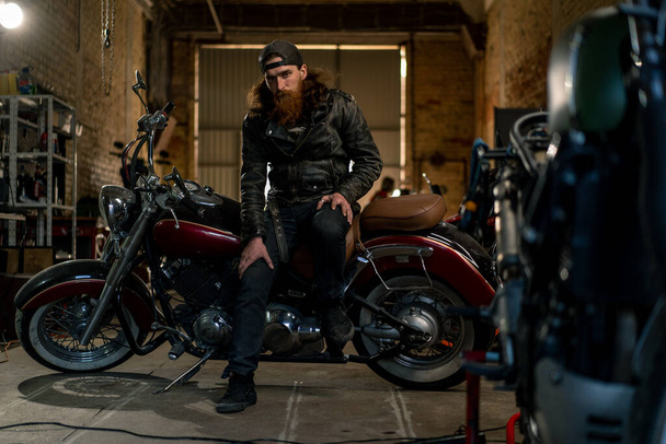 Δημιουργικό αυθεντικό εργαστήριο μοτοσικλέτας Garage βάναυση σοβαρά γενειοφόρος μηχανικός ποδηλάτης κάθεται στη μοτοσικλέτα - Φωτογραφία, εικόνα