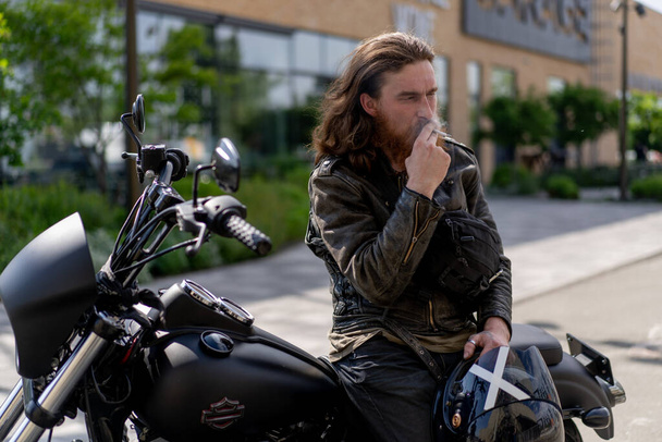 Brutaler, rotbärtiger Biker sitzt nach einem Rennen auf einem schwarzen Motorrad und raucht eine Zigarette nach Speed-Motorsport-Konzept - Foto, Bild