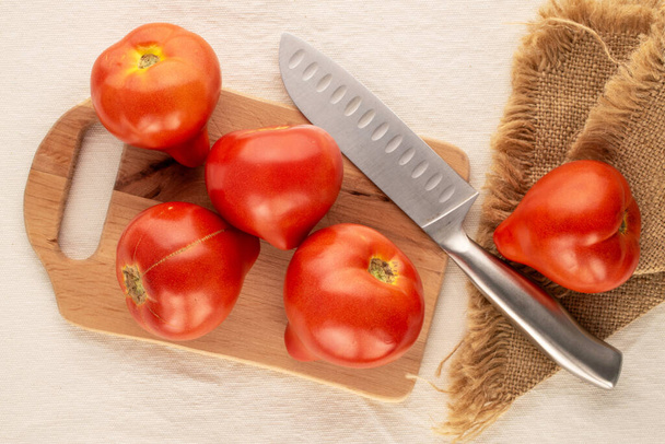 Several red tomatoes with wooden kitchen board and knife on linen cloth, macro, top view. - Foto, Bild