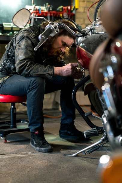 Δημιουργικό αυθεντικό εργαστήριο μοτοσικλέτας Garage κοκκινομάλλα γενειοφόρος μηχανικός ποδηλάτης επικεντρώνεται στην επισκευή μοτοσικλέτας - Φωτογραφία, εικόνα