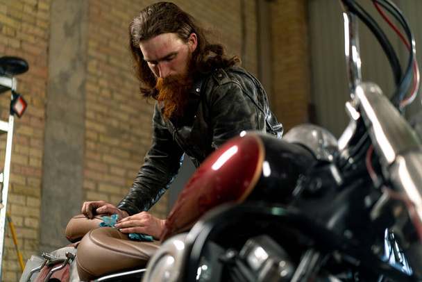 ラグが付いている美しいオートバイを拭く赤いひげ付きバイカーのメカニックの創造的な本物のオートバイの研修会 - 写真・画像