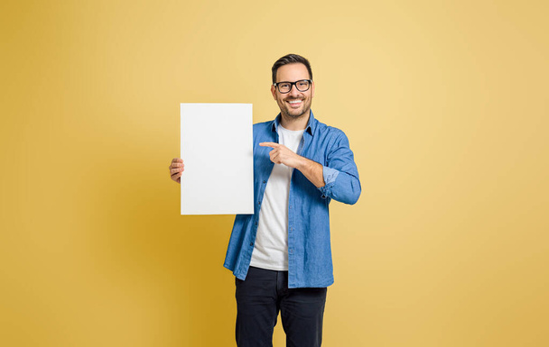 Lächelnder Geschäftsmann zeigt auf leeres weißes Plakat für Werbung vor gelbem Hintergrund - Foto, Bild