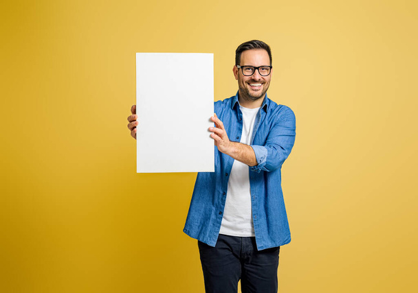 Happy male marketing manager tonen blanco witte poster voor reclame tegen gele achtergrond - Foto, afbeelding