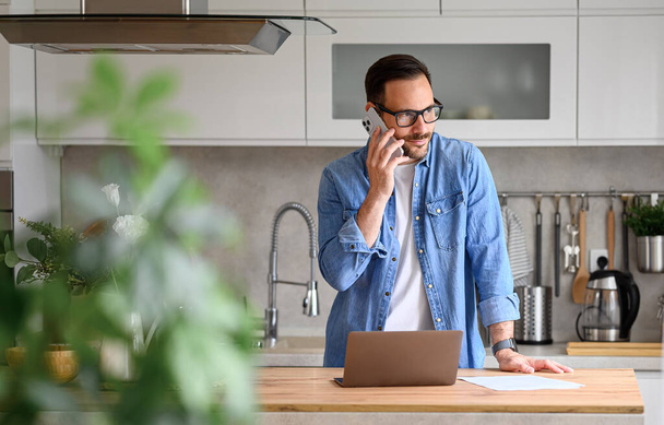 Professionnel masculin confiant parlant sur le téléphone intelligent tout en utilisant un ordinateur portable sur le comptoir de cuisine à la maison - Photo, image