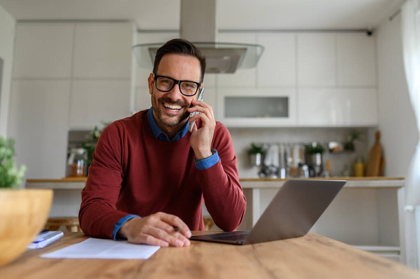 Portrait of smiling businessman talking over smart phone with laptop on desk at home office - Foto, Bild