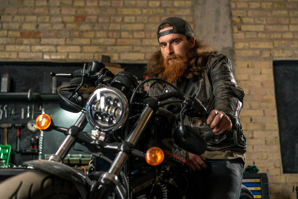 Créatif authentique atelier de moto garage portrait rousse grave barbu motard mécanicien assis sur la moto - Photo, image