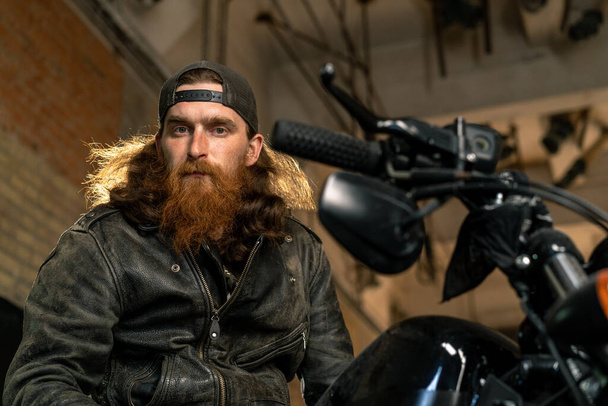 Yaratıcı otantik motosiklet atölyesi garajı portresi ciddi kızıl sakallı motosikletli tamirci motosiklette oturuyor. - Fotoğraf, Görsel