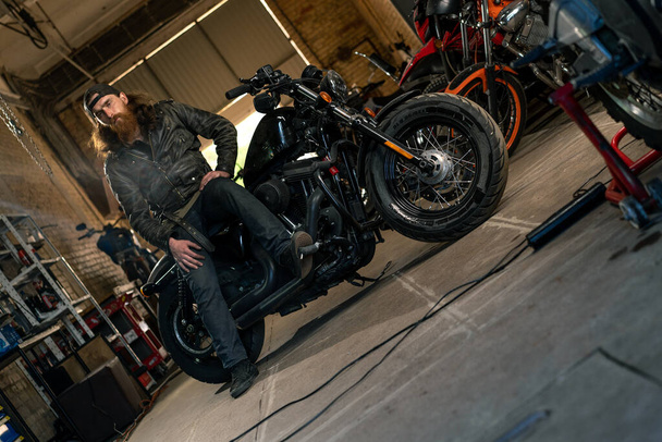 Креативный подлинный мотоцикл мастерская гараж серьезная рыжая бородатый мотоциклист механик, сидящий на мотоцикле - Фото, изображение