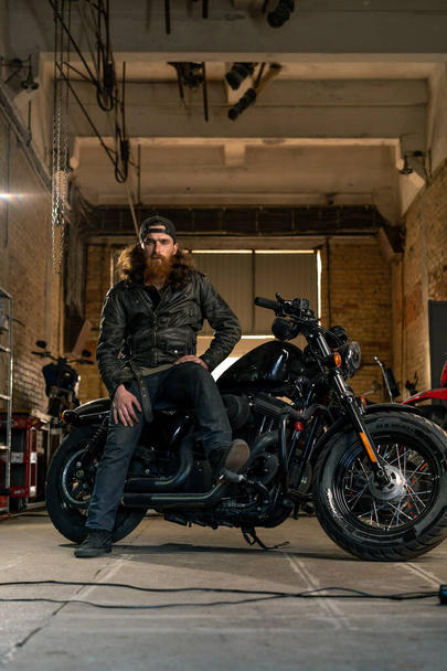 Creative authentic motorcycle workshop garage serious redhead bearded biker mechanic sitting on motorcycle - Fotografie, Obrázek