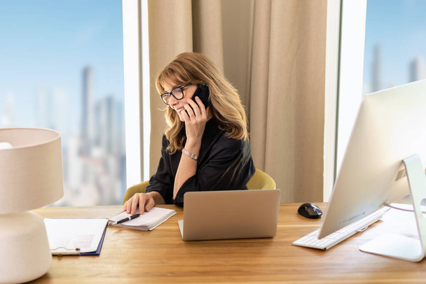 Shot of a mid aged woman sitting at desk and using mobile phone. Confident female wearing glasses and casual clothes and working in the office. - Photo, Image