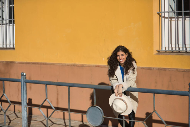 Young, beautiful, brunette woman with curly hair and beige coat and hat, leaning on a railing looking at camera smiling and happy. Concept beauty, fashion, autumn, winter, cold. - Photo, image