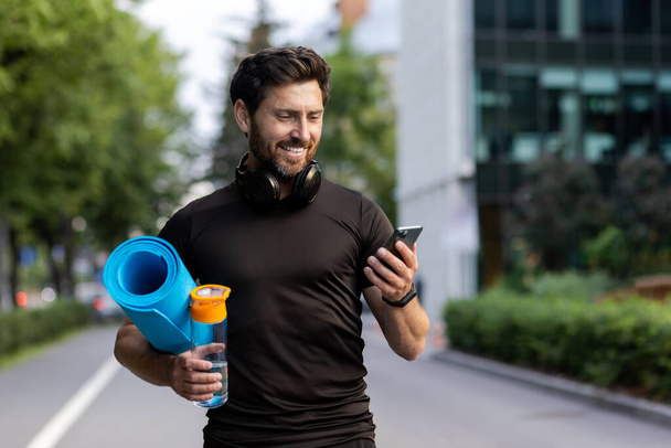 Portrait of mature successful adult athlete, bearded man going to workout, coach holding fitness mat and phone, browsing online app for joint physical training. - Foto, Imagen