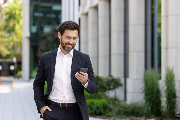 Mature adult male boss walking outside office building, successful businessman typing message and browsing online pages happily, investor in business suit with smartphone. - Photo, Image