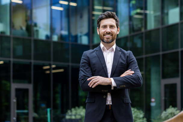 Portrait of mature adult businessman, senior boss with beard smiling and looking at camera, experienced and successful man in business suit outside office building with crossed arms. - Photo, Image
