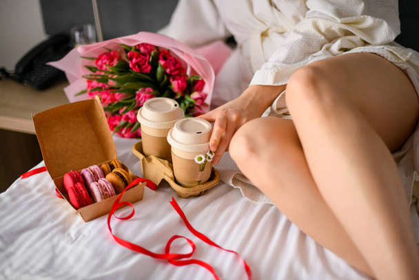 Tray with fresh cup of coffee and macarons dessert standing near womans legs. Breakfast in bed concept. Bouquet with colorful tulips lying on bed. Flovers delivery concept - Photo, Image