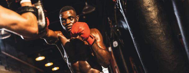 Boxer africain frappant à l'entraîneur ou entraîneur portant des mitaines de poinçonnage comme équipement d'entraînement de sac de boxe dans la salle de gym. Entraînement de force et d'endurance pour un match de boxe professionnel. Éperon - Photo, image