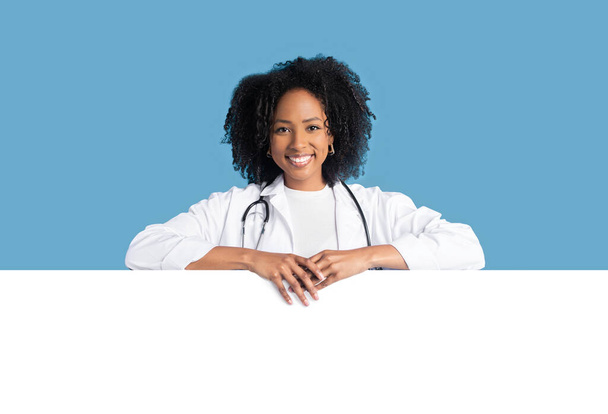 Happy young african american curly woman doctor therapist in white coat with big banner with blank space, isolated on blue background, studio. Ad and offer, medicine service, health care - Photo, Image