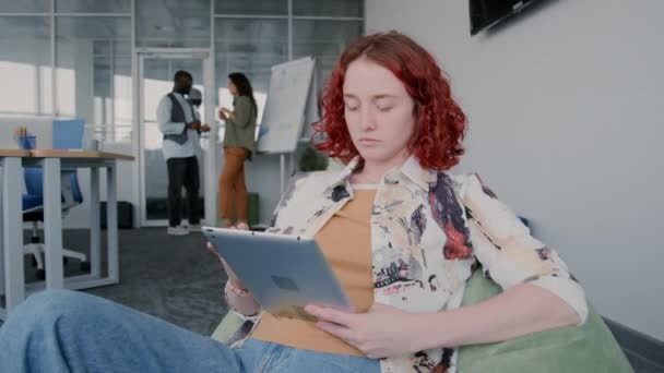 Середній портрет молодої жінки з червоним кучерявим волоссям, що сидить на стільці для мішків з квасолею, що працює на цифровому планшеті
 - Кадри, відео