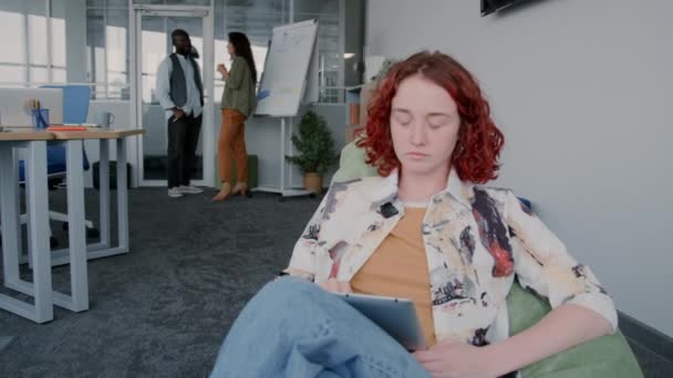 Kırmızı kıvırcık saçlı genç bir kadının portresi, gündelik giysiler giymiş, fasulye koltukta oturmuş, modern ofiste dijital tablet üzerinde çalışıyor. - Video, Çekim