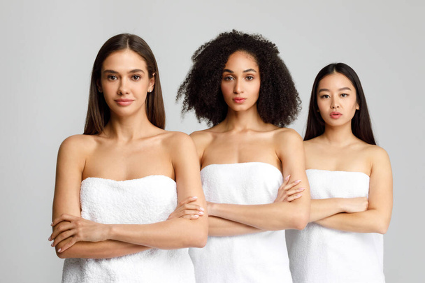 Schöne schwarze, kaukasische und asiatische Frauen posieren mit verschränkten Armen in weißen Badetüchern, stehen Schlange vor grauem Hintergrund, Studioaufnahme, Banner - Foto, Bild