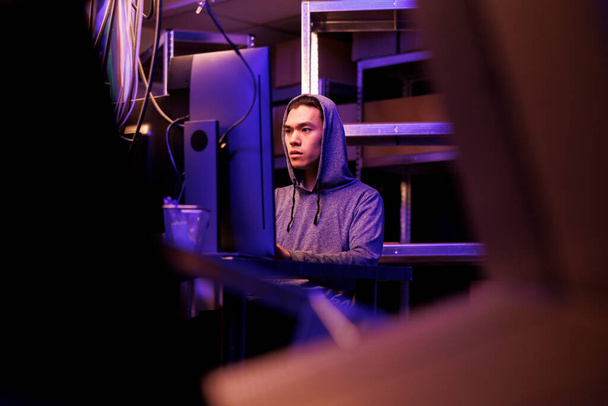 Asian hacker working on ransomware to break into government server and steal sensitive data. Young man in hood coding illegal malicious software and programming spyware at night - Photo, image