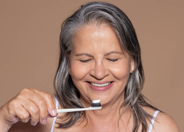 Usmívající se evropská starší šedovlasá žena si kartáčkem čistí zuby, užívá si denní rutiny, izolovaná na pozadí béžového studia, zblízka. Higiena, zubní péče, reklama, nabídka - Fotografie, Obrázek