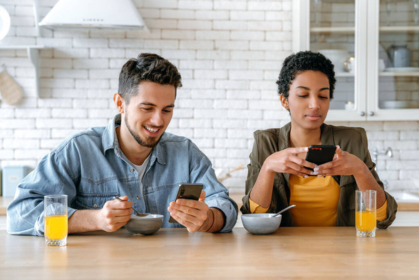 Frühstück. Verliebtes multirassisches Paar, afrikanisch-amerikanische Frau und kaukasischer Mann, die ihr Mobiltelefon während des gemeinsamen Frühstücks zu Hause benutzen, Online-Videos oder Nachrichten ansehen, Nachrichten verschicken - Foto, Bild
