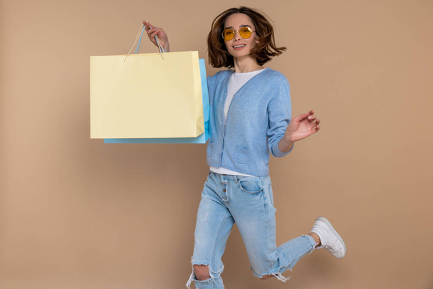 Overjoyed woman in casual clothing jumping up high with shopping bags in hands isolated over beige background. - Φωτογραφία, εικόνα