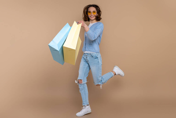 Happy joyful woman in casual clothing running jumping with shopping bags isolated over beige background. - Фото, изображение