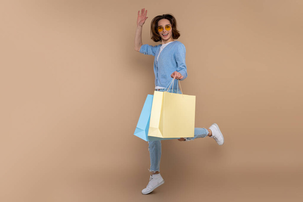 Overjoyed woman shopaholic in casual clothing jumping with shopping bag purchases isolated over beige background. - Valokuva, kuva